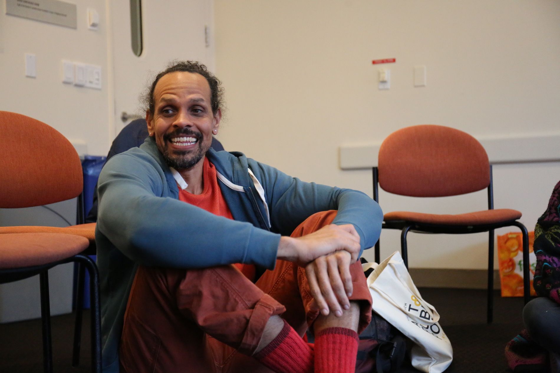 Ross Gay sits on the floor holding a paper with his knees up to his chest smiling at the camera